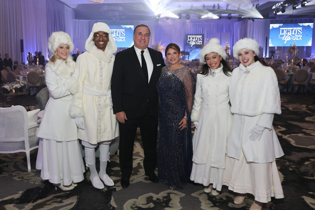 Two guests stand with performers in white winter costumes at the gala.